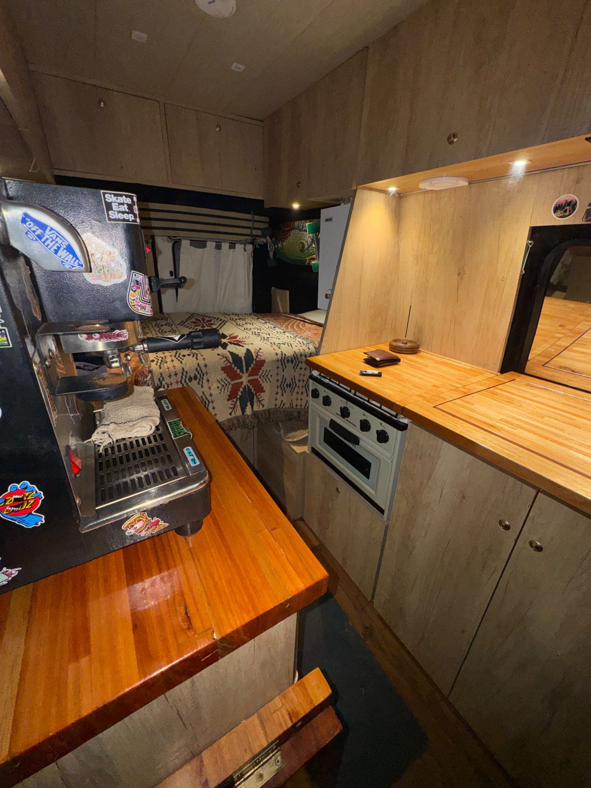 Interior de la van con la cafetera, la mesada de madera y la cama al fondo
